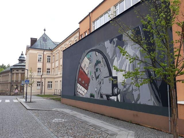 Streetart na budově gymnázia, pohled na část komplexu školy s patrnými zálivy pro parkování vozidel mezi alejí nově vysazených lip