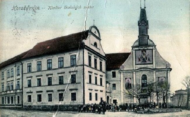 Kolorovaná pohlednice klášterního kostela kolem roku 1910. Budova školy v popředí je již po nástavbě třetího patra.